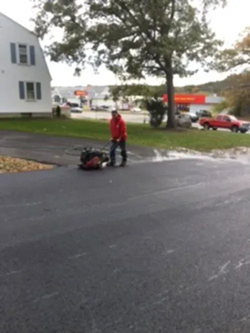 Man operating a leaf blower on wet asphalt. Building and cars visible in the background. Overcast sky.