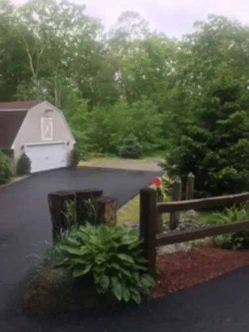 Barn with a white garage door, asphalt driveway, wooden fence, lush green foliage. Overcast sky.