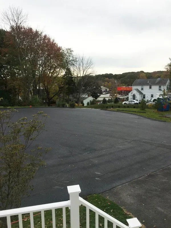 Newly paved asphalt lot with a white railing in the foreground, houses and trees in the distance under an overcast sky.