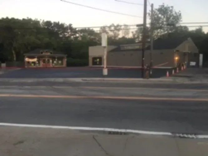 Buildings behind a road; one with an awning, the other with a chimney. Gray asphalt, trees, and sky are visible.