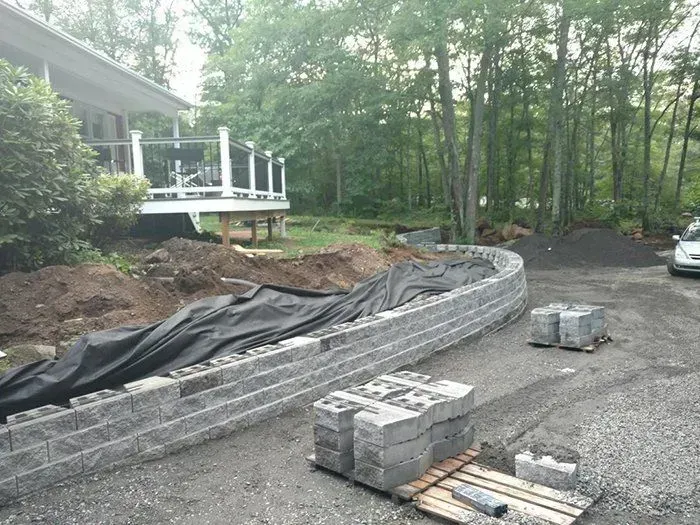 Construction site with a retaining wall being built beside a house, gravel driveway, and a small pile of dirt.