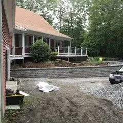 Red house with porch and retaining wall, car parked in gravel driveway.