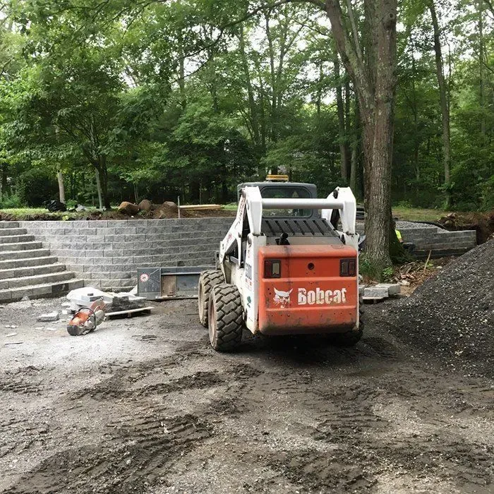 Bobcat skid steer working on a stone retaining wall in a wooded area.