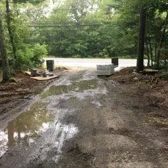Dirt road with puddles, leading to a paved road, flanked by trees and greenery.