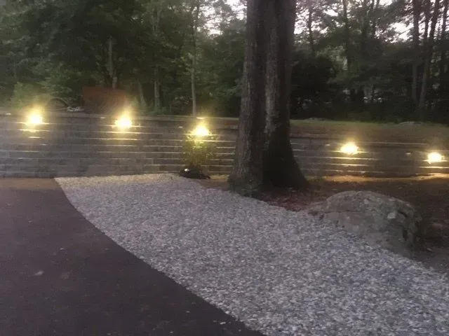 Outdoor scene with lit steps, gravel path, tree, and surrounding greenery. Evening.