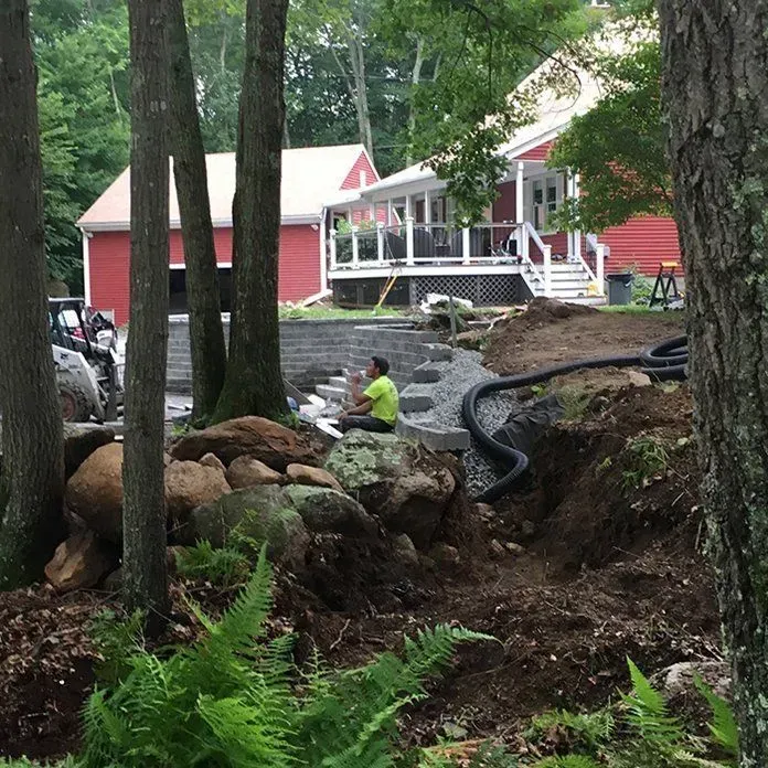 Construction site: man working on stone steps near a red house and retaining wall, trees in foreground.