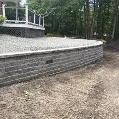 Gray retaining wall with gravel surface, next to a deck and wooded area.