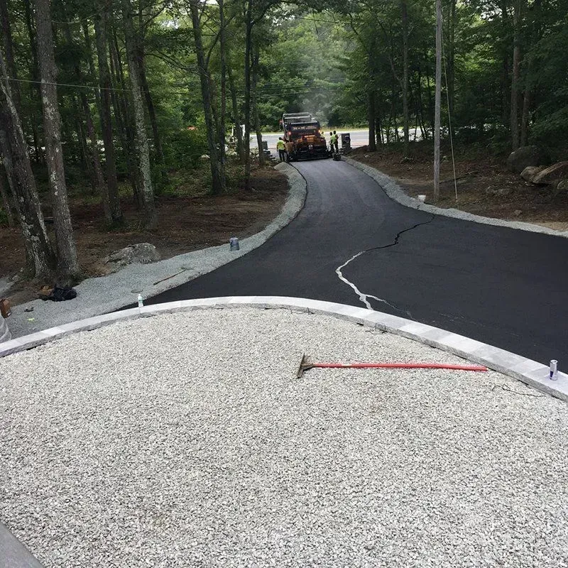 Asphalt paving a curved driveway, stone base in foreground, construction equipment in the distance.
