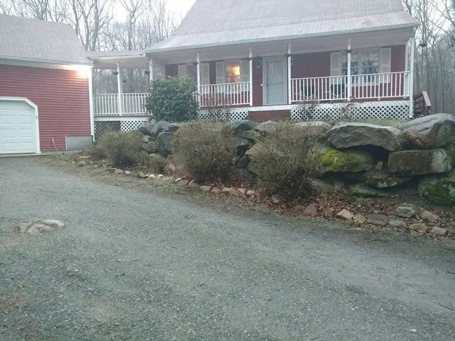 Red house with porch, stone retaining wall, and gravel driveway. Barn to the left.