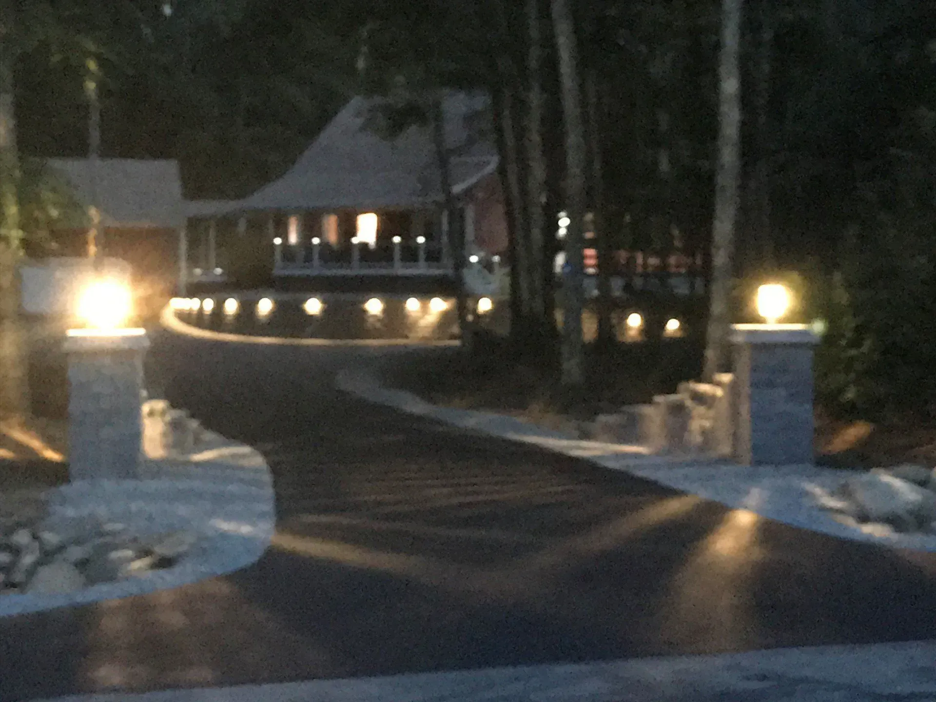 Dark asphalt driveway leading to a house, illuminated by lights on pillars and along the house's foundation.