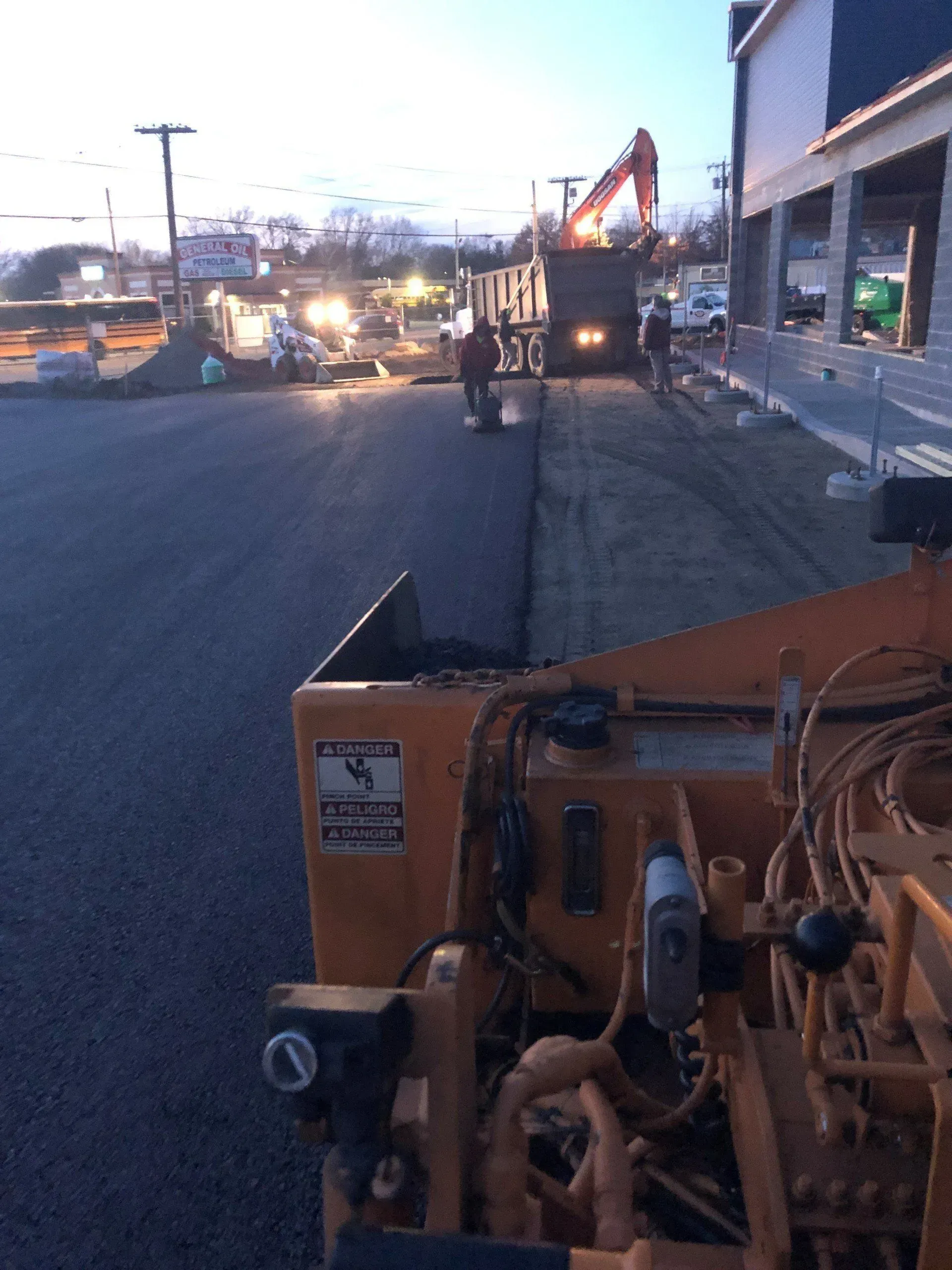 Asphalt paving in progress; workers, machinery, and trucks at dusk.