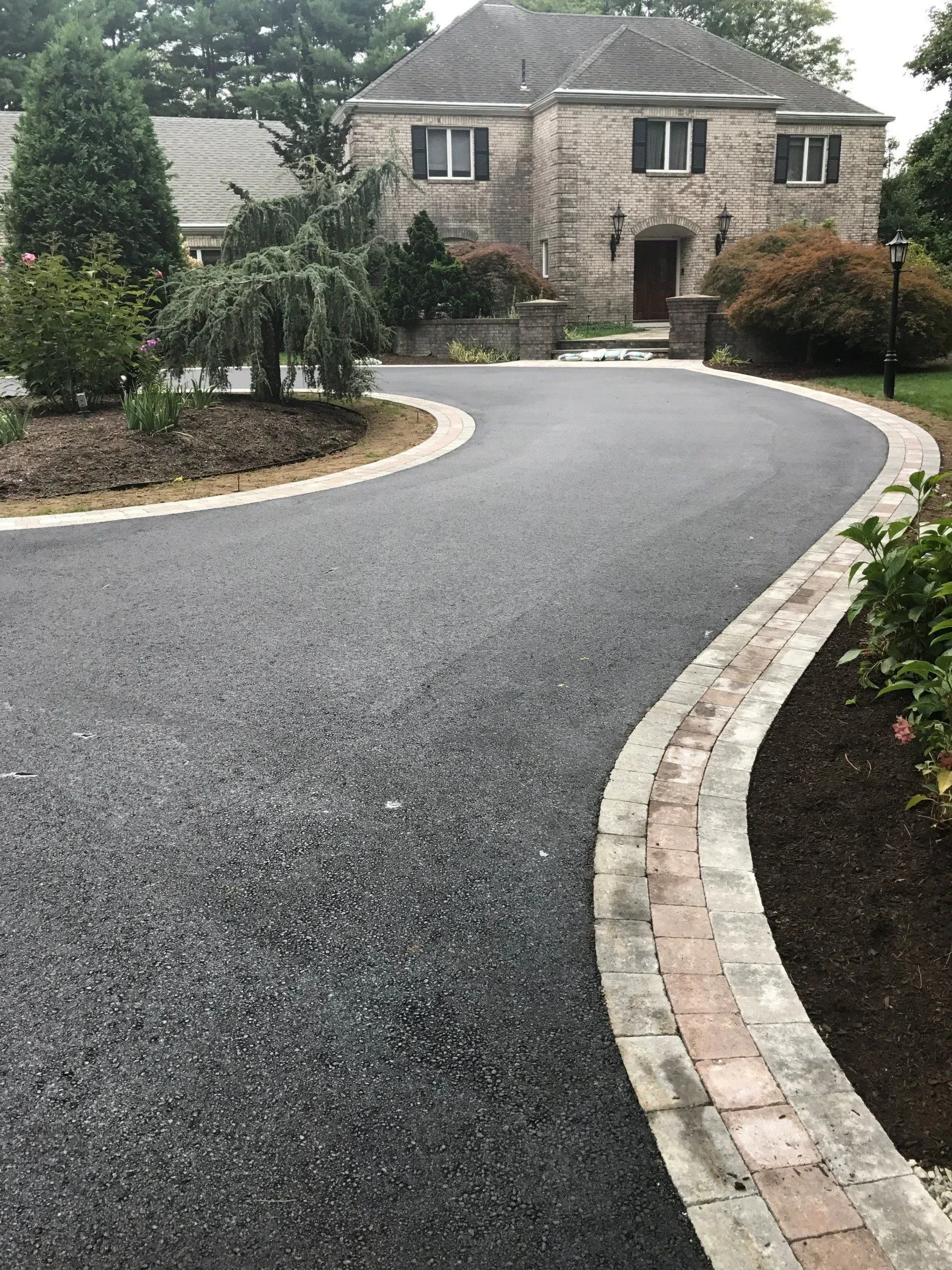 Asphalt driveway curves toward a brick and stone house, edged with brick and concrete.
