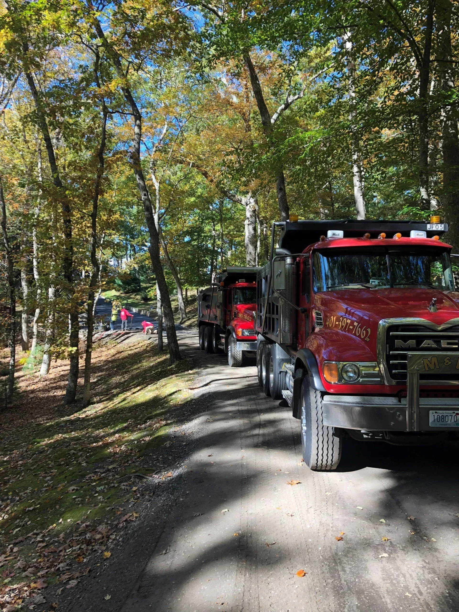 Two red dump trucks parked on a dirt road in a wooded area with fall foliage.