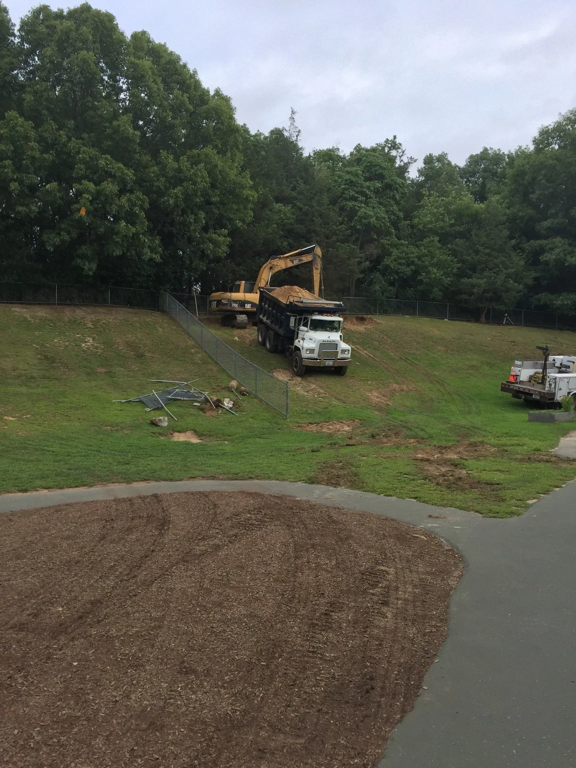 Excavator loading a dump truck on a grassy hill; trees in the background, cloudy sky.