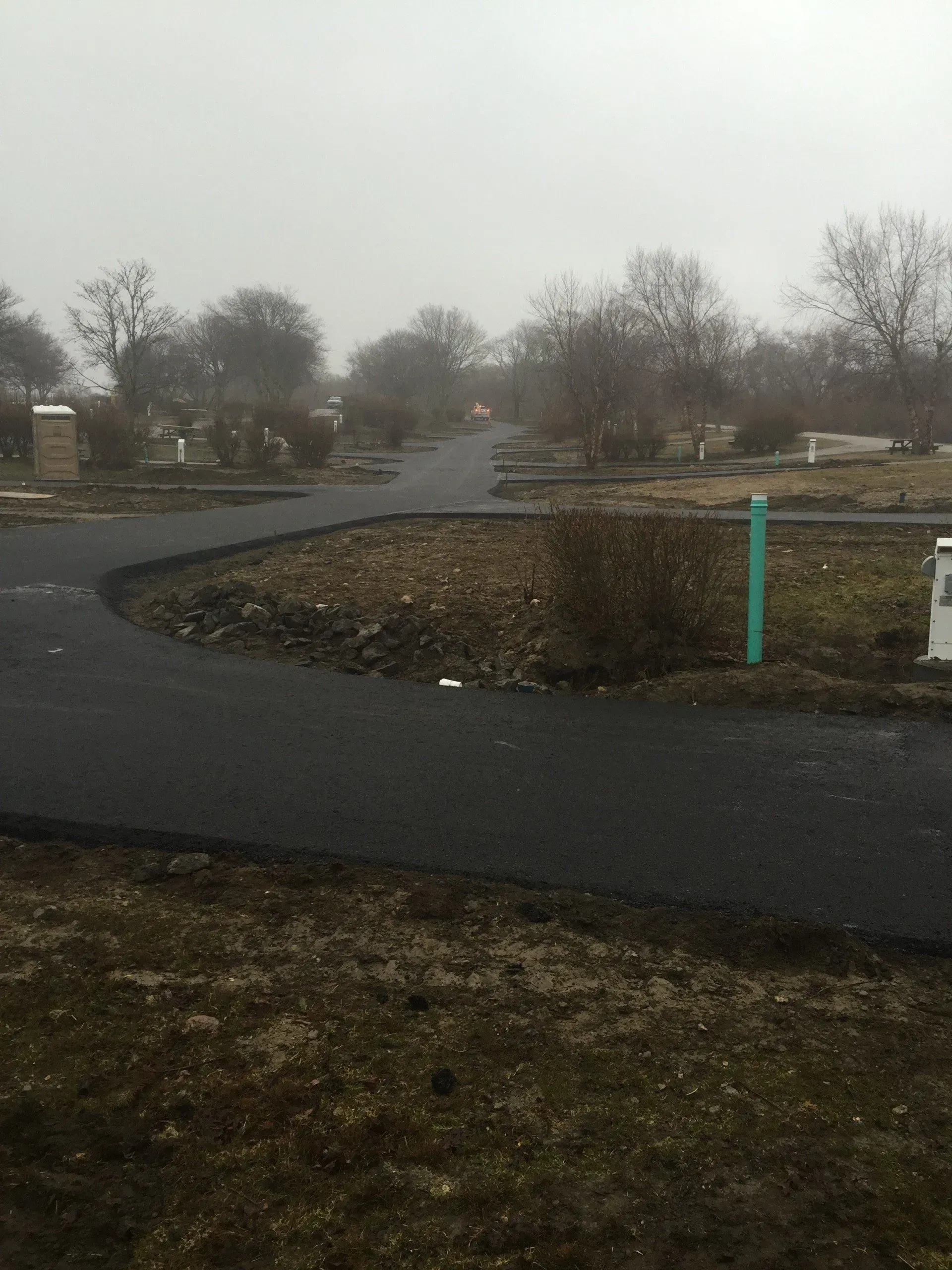 Asphalt road curves through a foggy cemetery with bare trees and small green pipe.