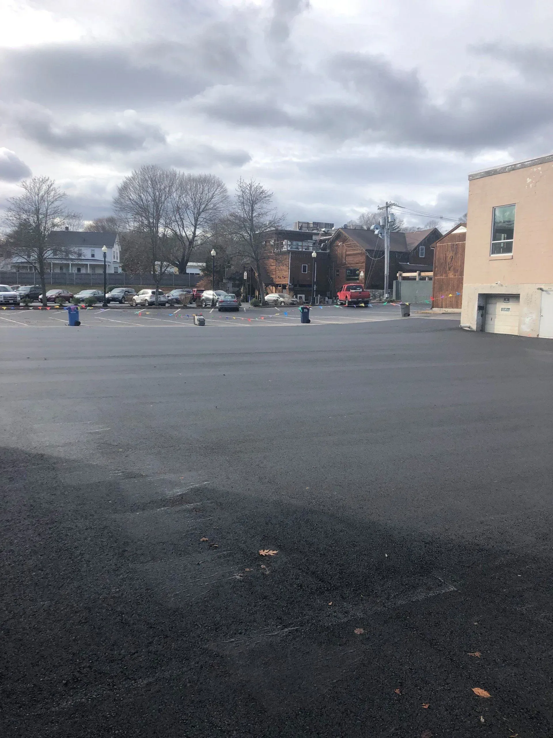An empty asphalt parking lot with parked cars in the background under a cloudy sky.