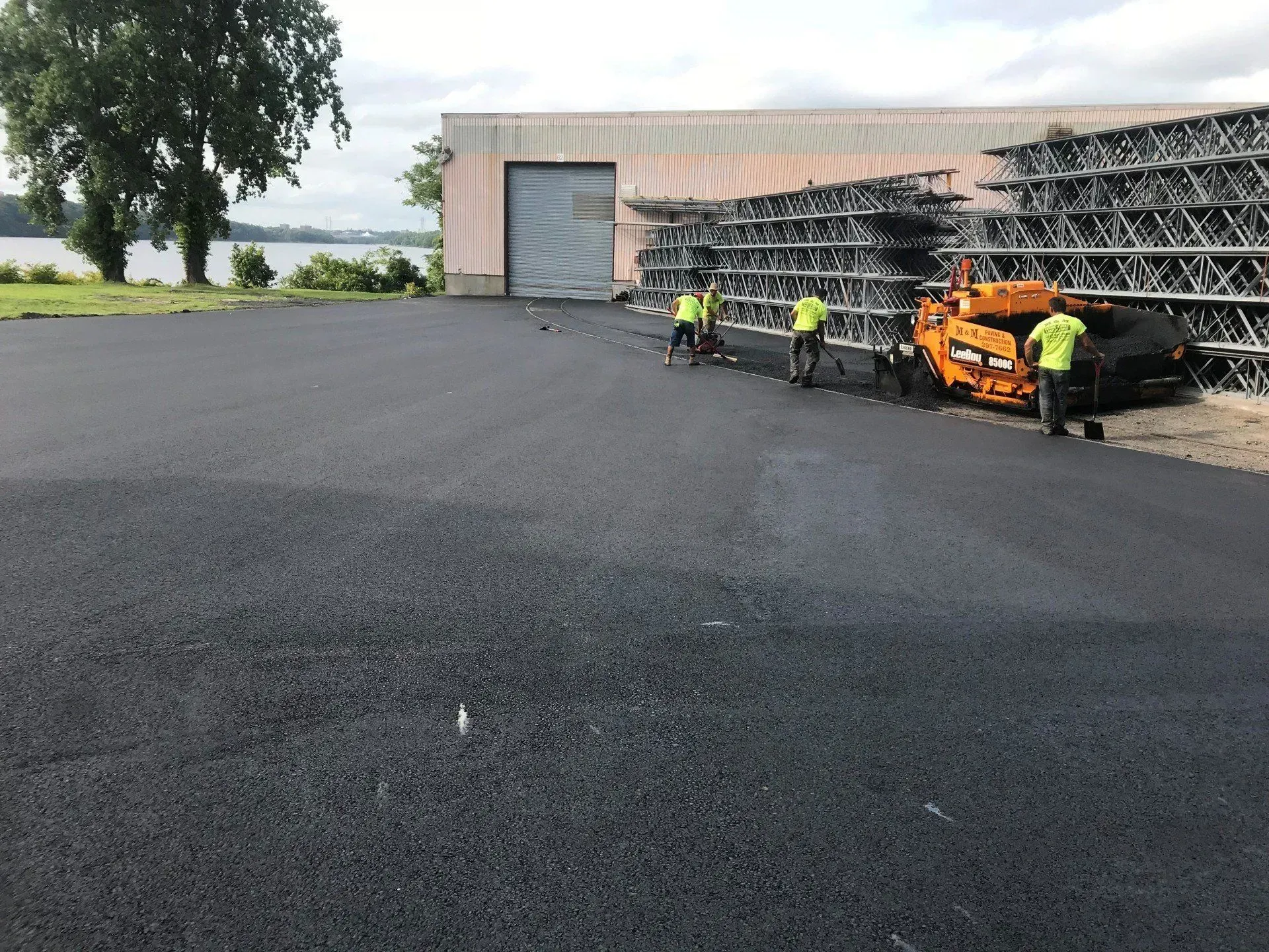 Asphalt paving in progress; workers in yellow vests operate machinery near a large warehouse and body of water.