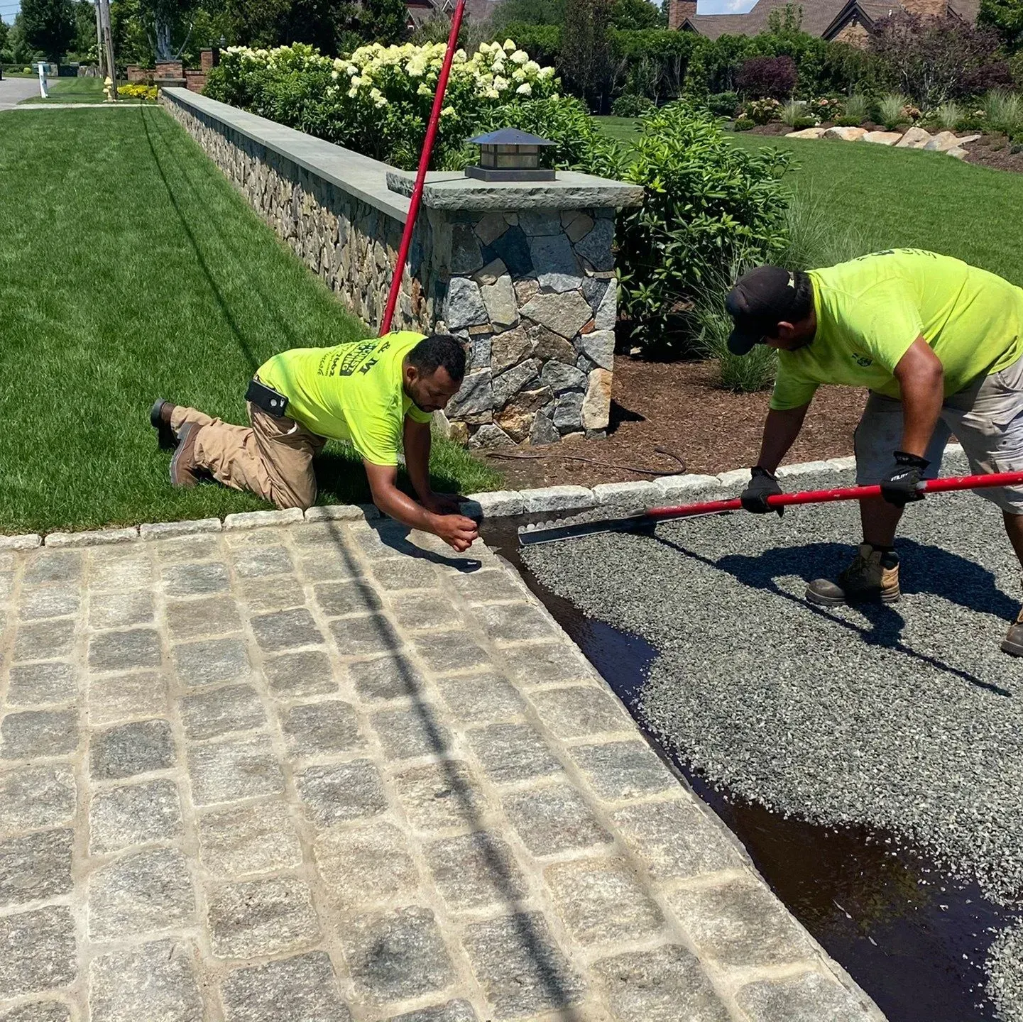 Two workers in green shirts install gravel next to a stone walkway and wall, outdoors.