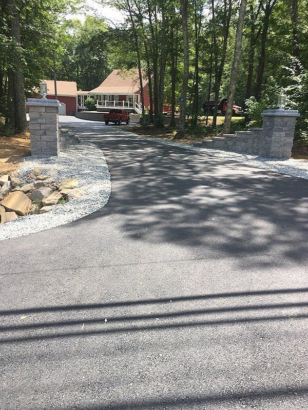 Asphalt driveway leading to a red house with stone entrance pillars. Trees line the sides.