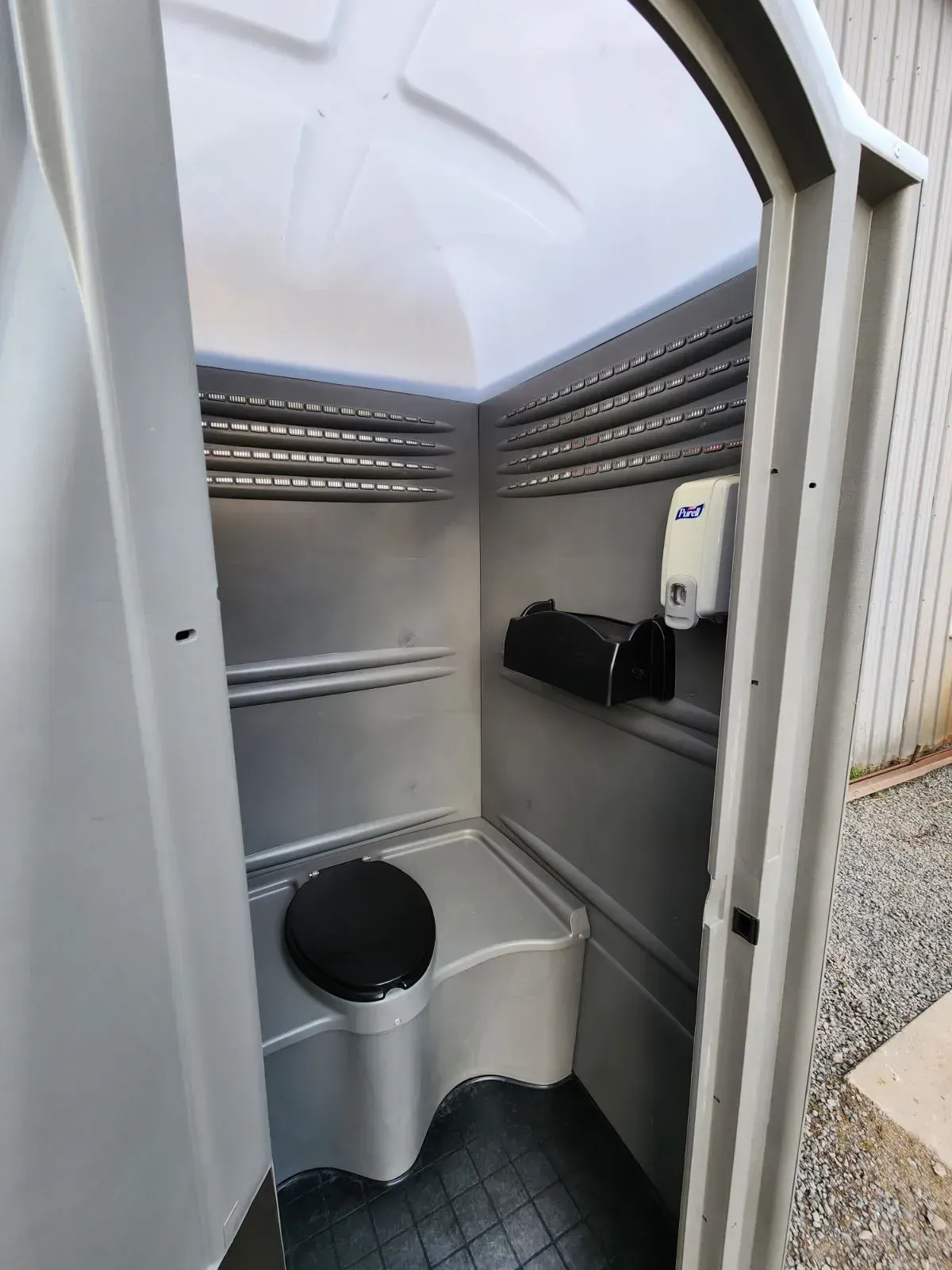Interior view of a portable toilet. Gray walls, black toilet seat, soap dispenser, and paper towel holder are visible.