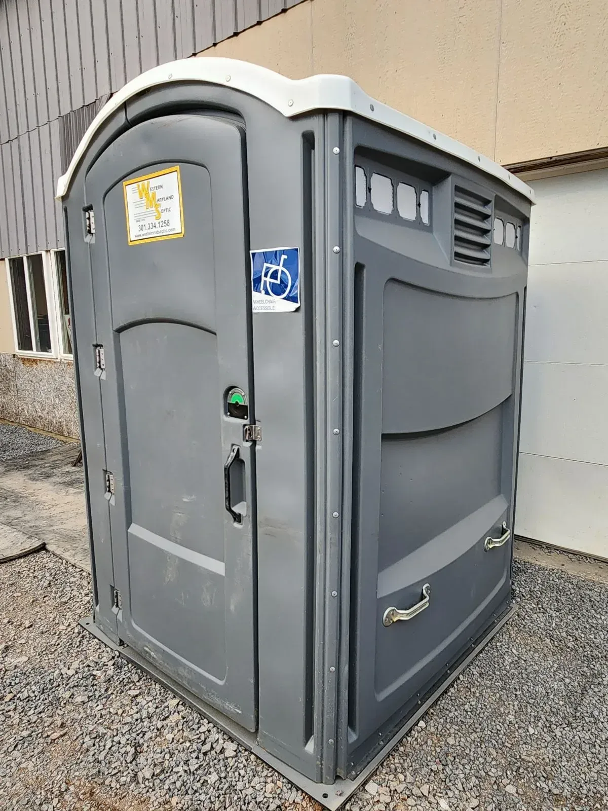 Gray portable toilet with accessible door sticker, outdoors on gravel.