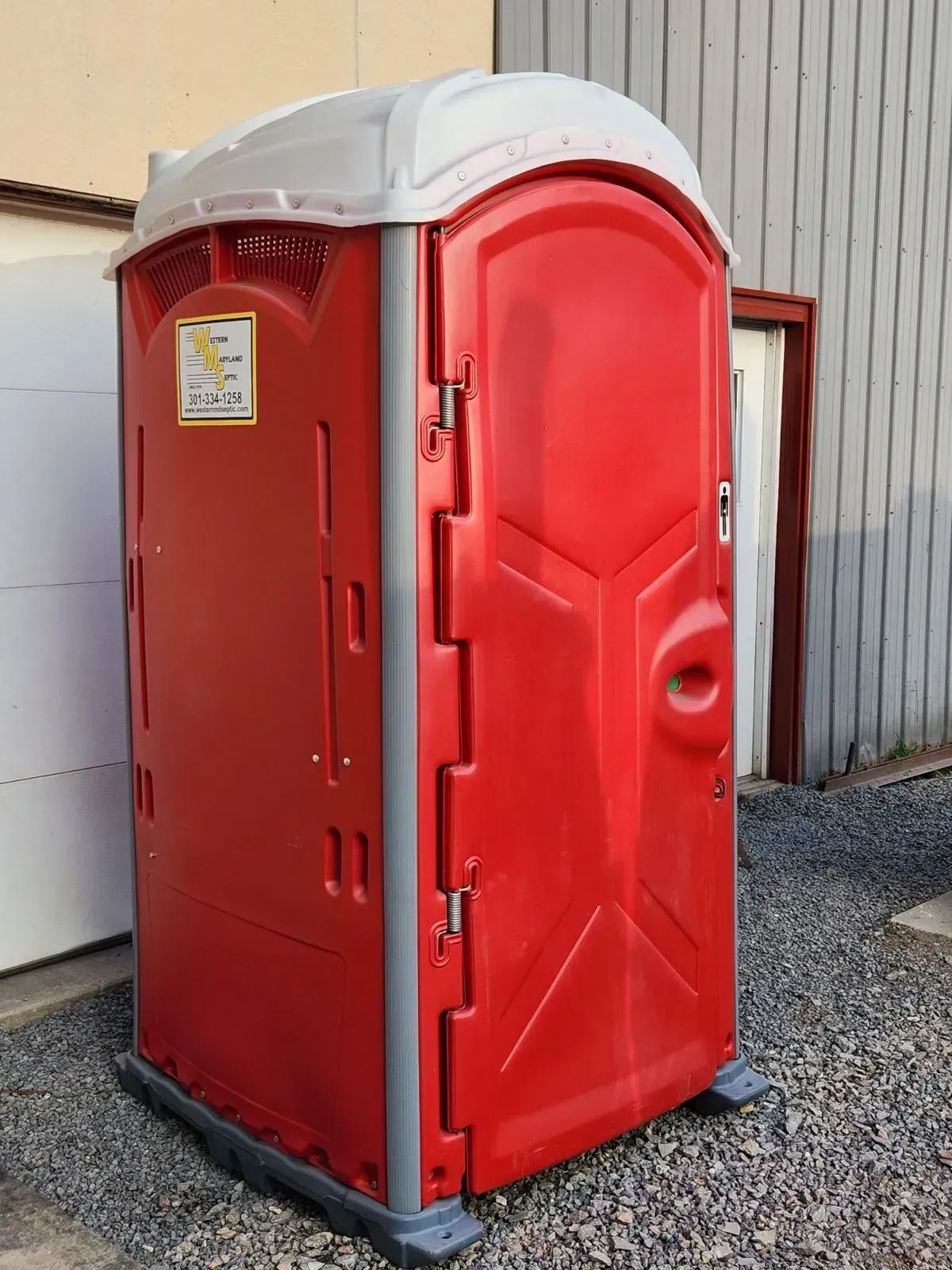 Red and white portable toilet on a gravel surface next to a building.