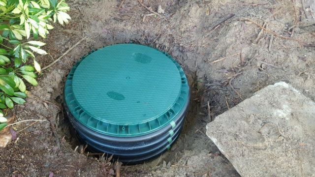Green septic tank lid in a hole in the ground, next to a stone.