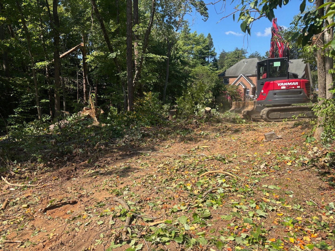 Red excavator clearing a dirt path beside trees near a house on a sunny day