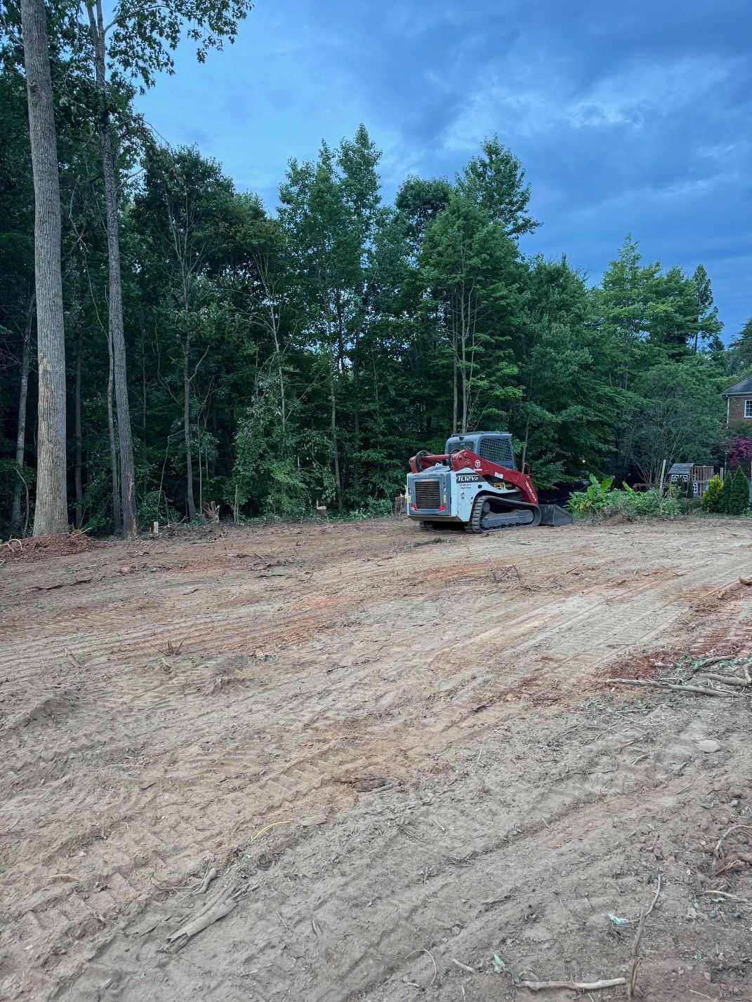 Cleared dirt lot by trees with a red skid steer near a wooded edge under a blue sky
