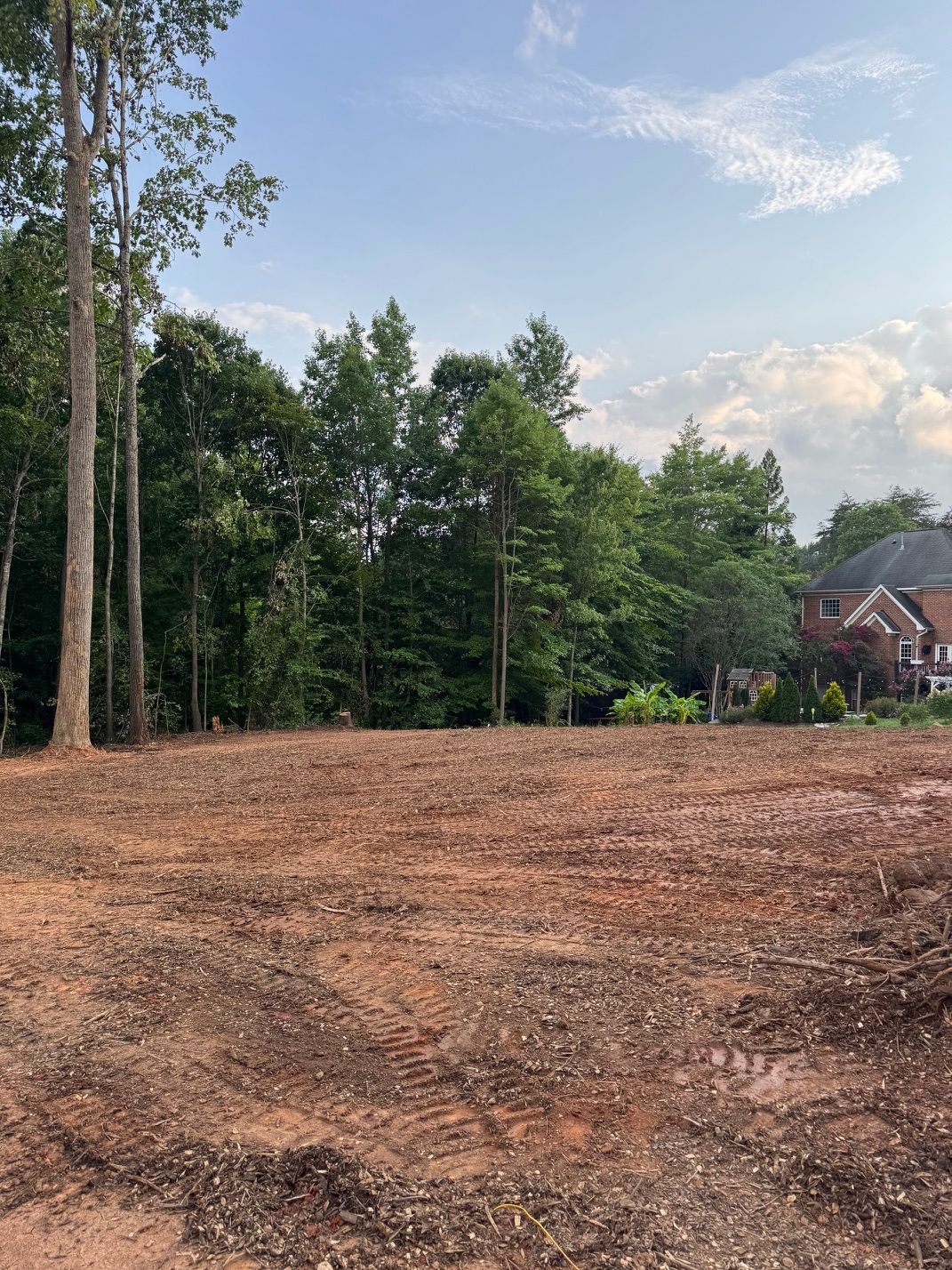 Cleared dirt lot beside tall trees and a house under a partly cloudy sky