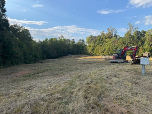 Cleared grassy field bordered by trees, with a red excavator and a white utility box on the right