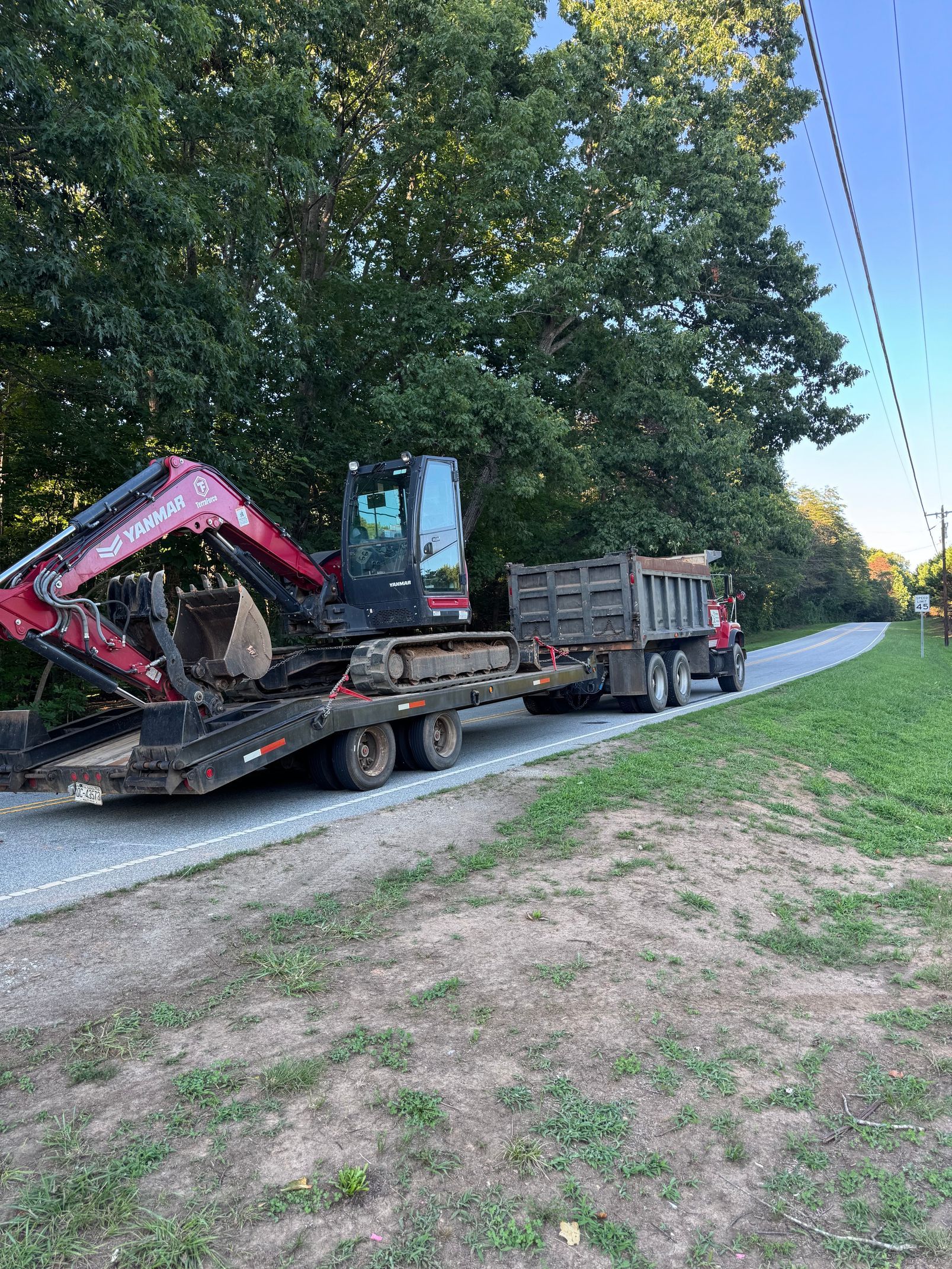 Flatbed truck hauling a red excavator on a gravel road beside trees and power lines
