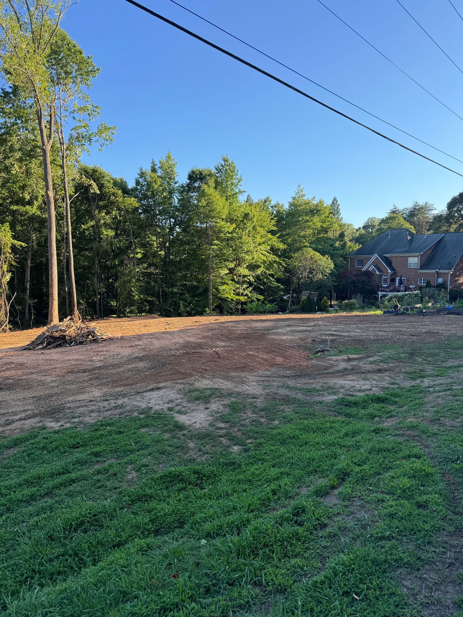 Cleared dirt lot beside a wooded area and houses under a blue sky, with power lines overhead
