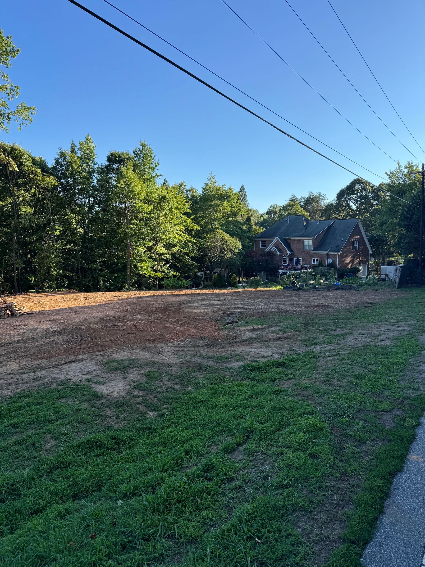Grassy roadside lot beside trees and power lines, with houses in the distance under a clear blue sky.