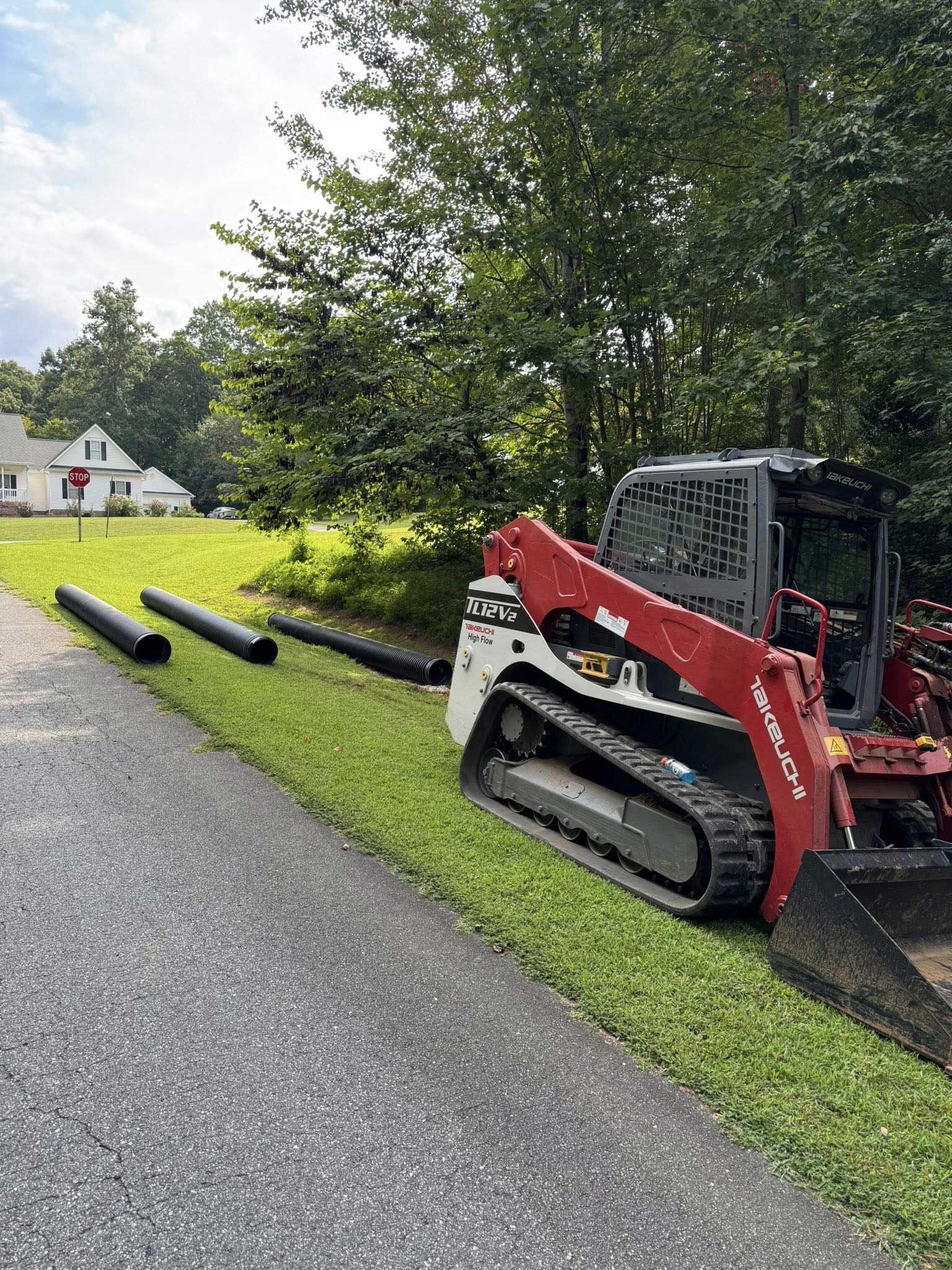 Red and white skid steer next to a roadside with black pipes and a grassy edge. A house is in the background.