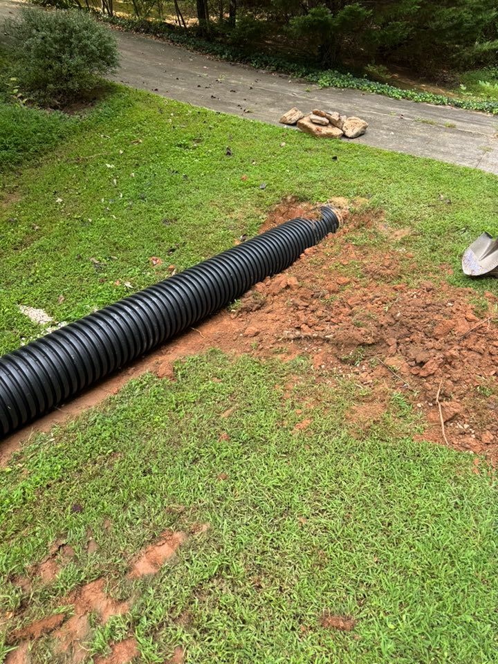 Black corrugated drainage pipe laid across a grassy lawn, heading toward a dirt patch by a driveway.