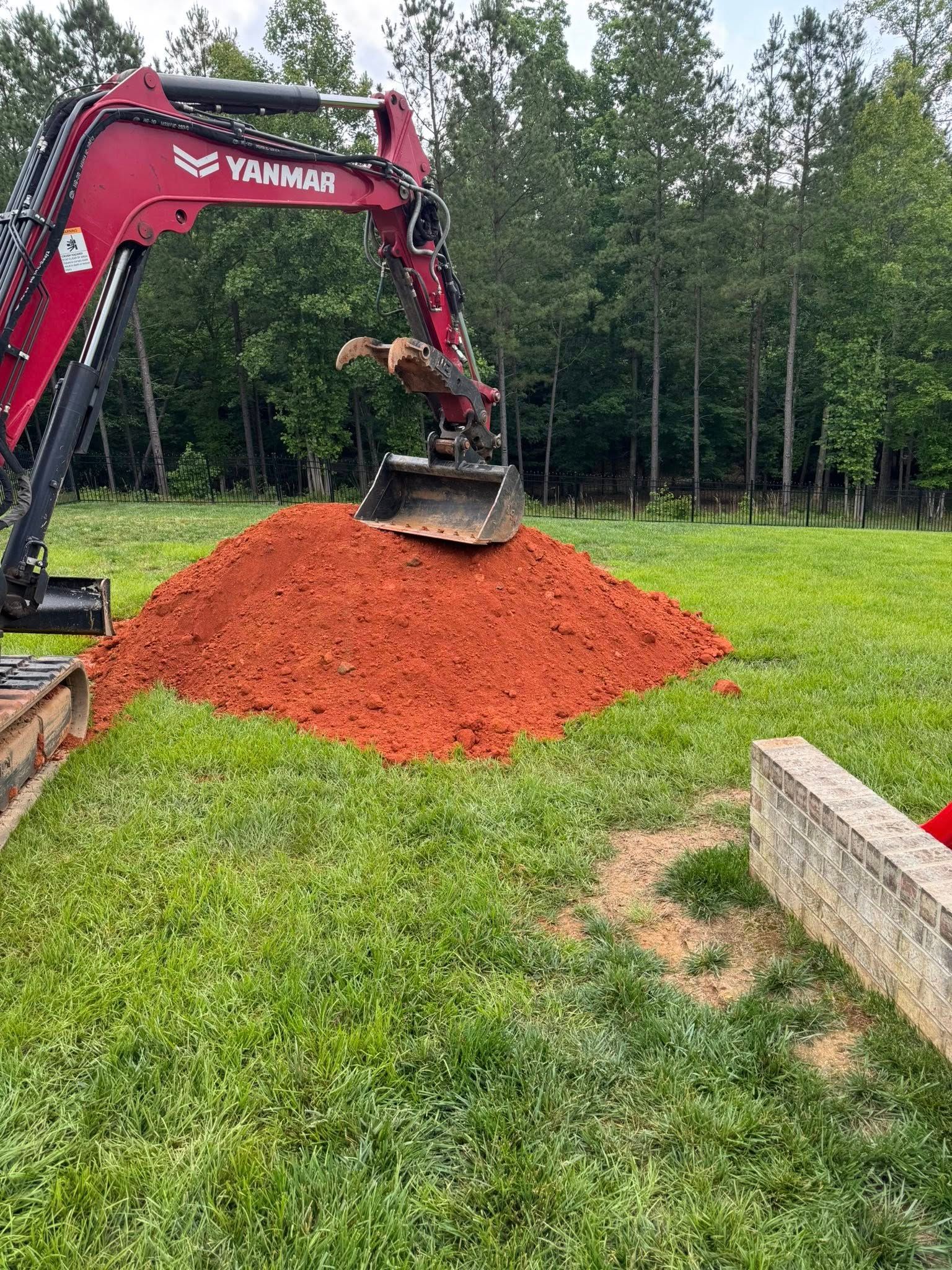 Red excavator moving red dirt on a green lawn near trees.