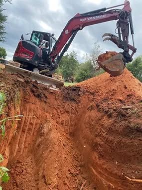 Excavator digging in red soil, creating a trench. Dark red machine with operator.