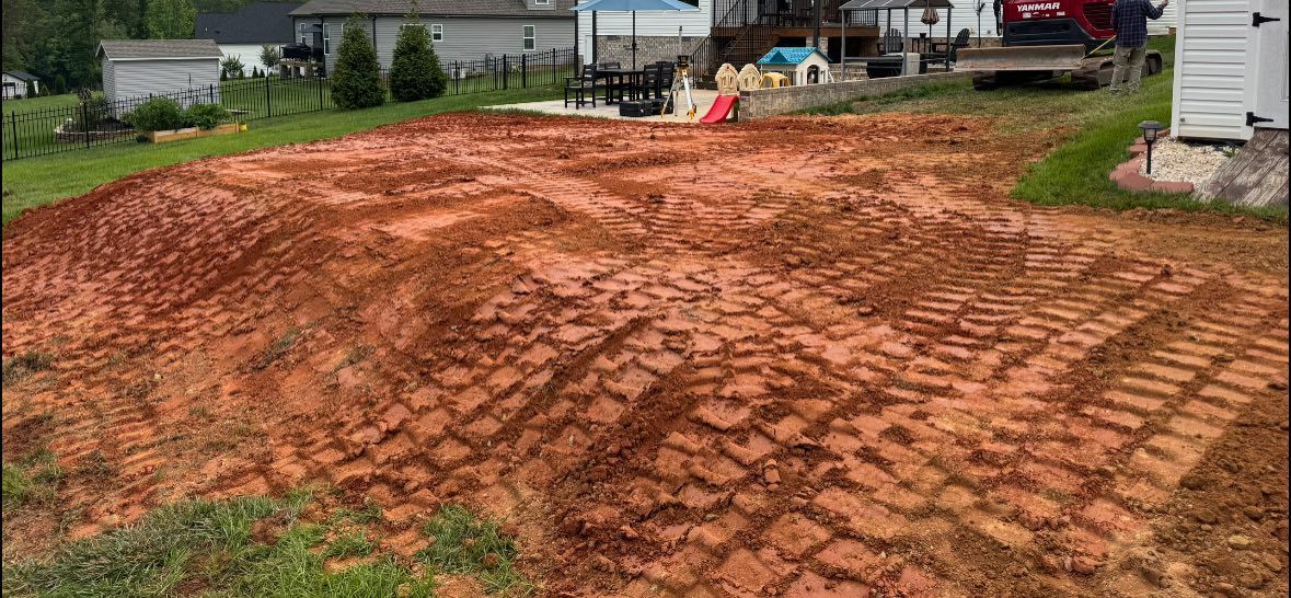 A freshly tilled backyard, with red soil and tractor tracks. Houses and grass are in the background.