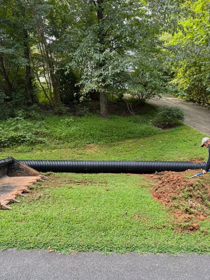 A black corrugated pipe stretches across grass, where dirt has been dug up. A person works on the right.