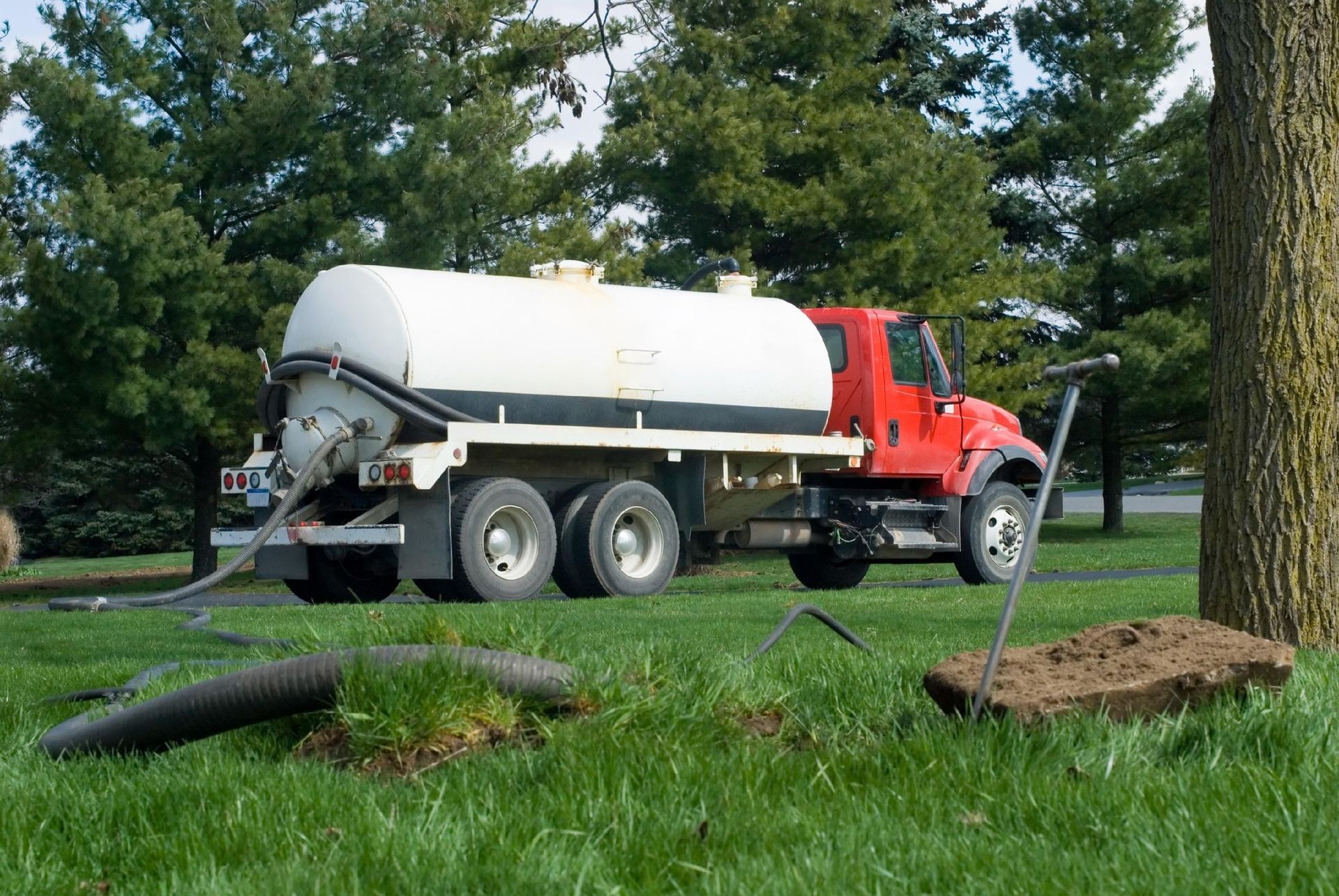 Red septic truck parked on grass, hose connected to ground. Trees in background.