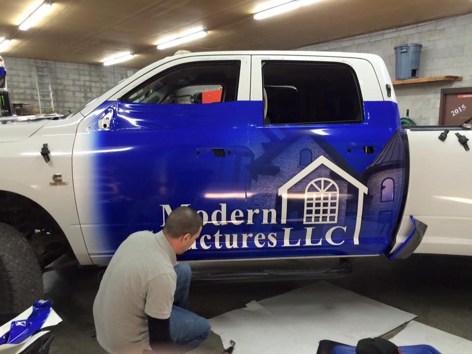 A man is wrapping a blue and white truck with modern pictures llc written on it