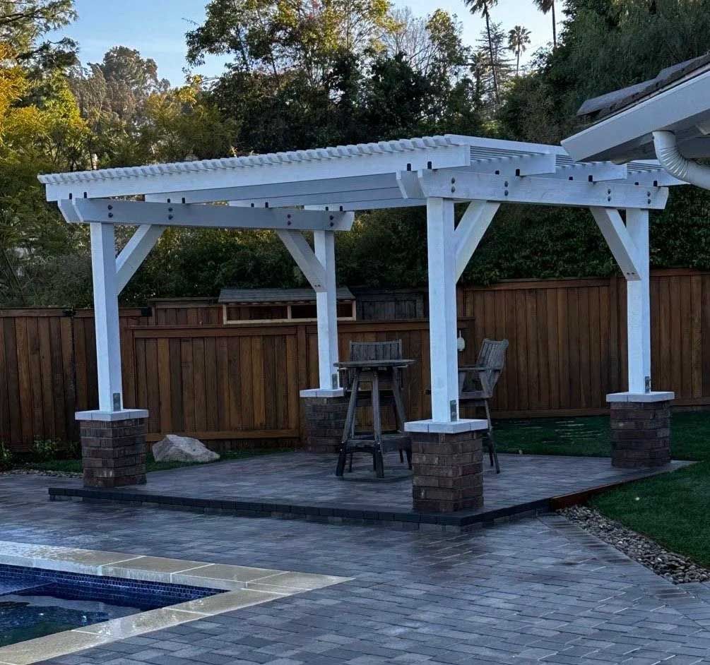 White pergola with a stone base over a patio with outdoor furniture, beside a pool and wooden fence.