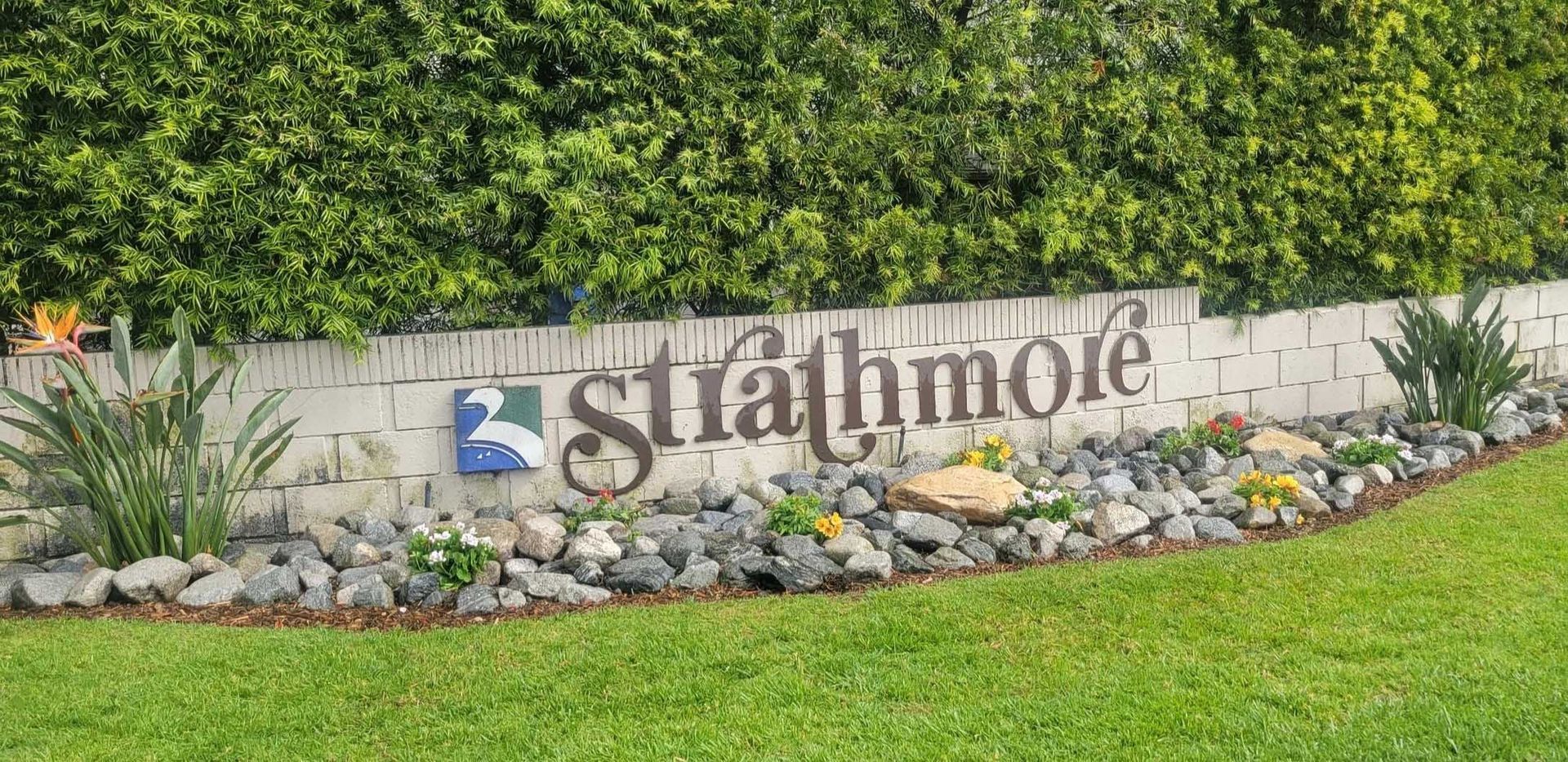 Sign for Strathmoie neighborhood with a brick wall, green foliage backdrop, and a logo featuring a bird.