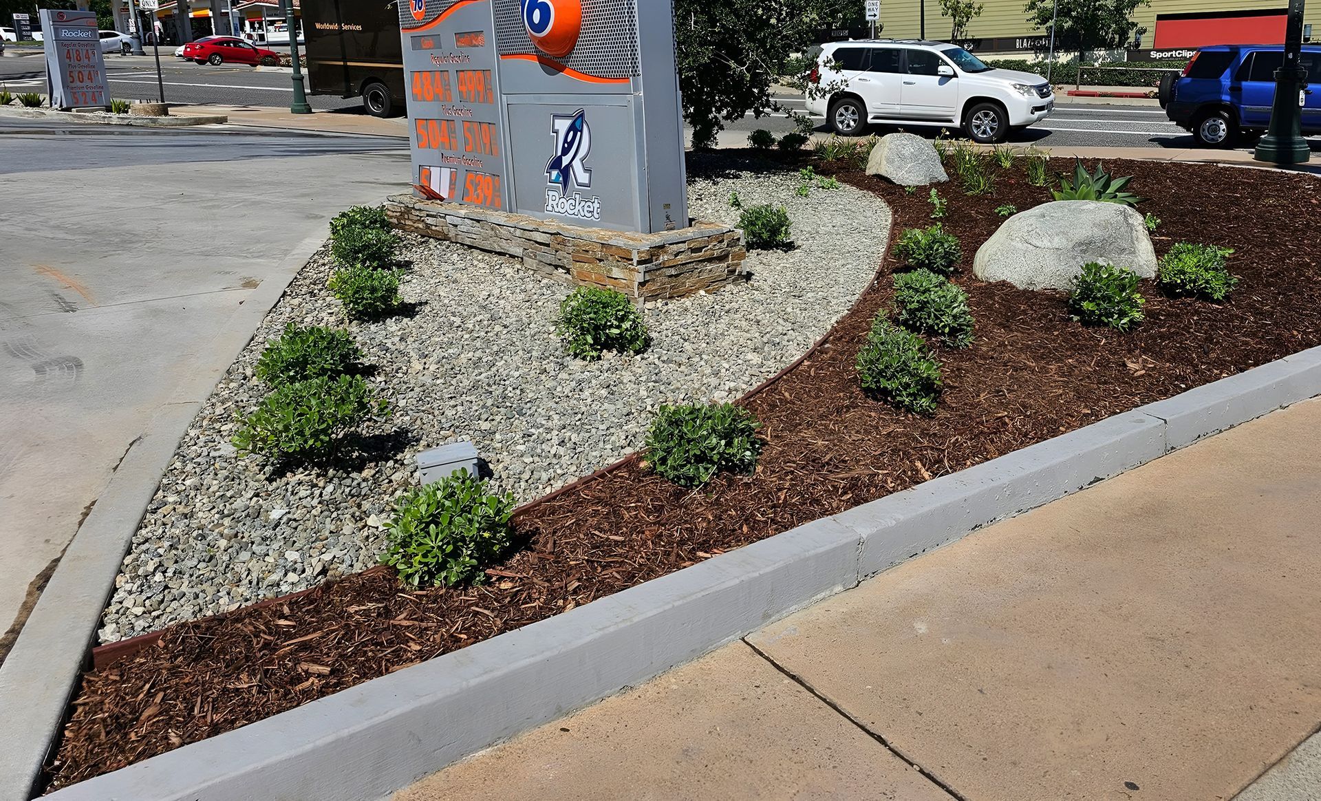 A small triangular garden bed with green bushes, rocks, and mulch, bordered by concrete, next to a sidewalk and a sign.