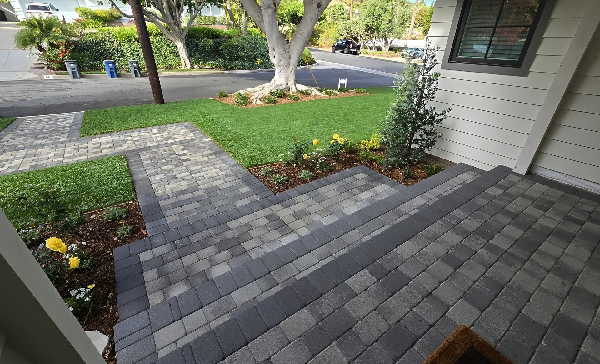 Brick steps leading up to a house entrance with lush landscaping, including a central tree and surrounding lawn.