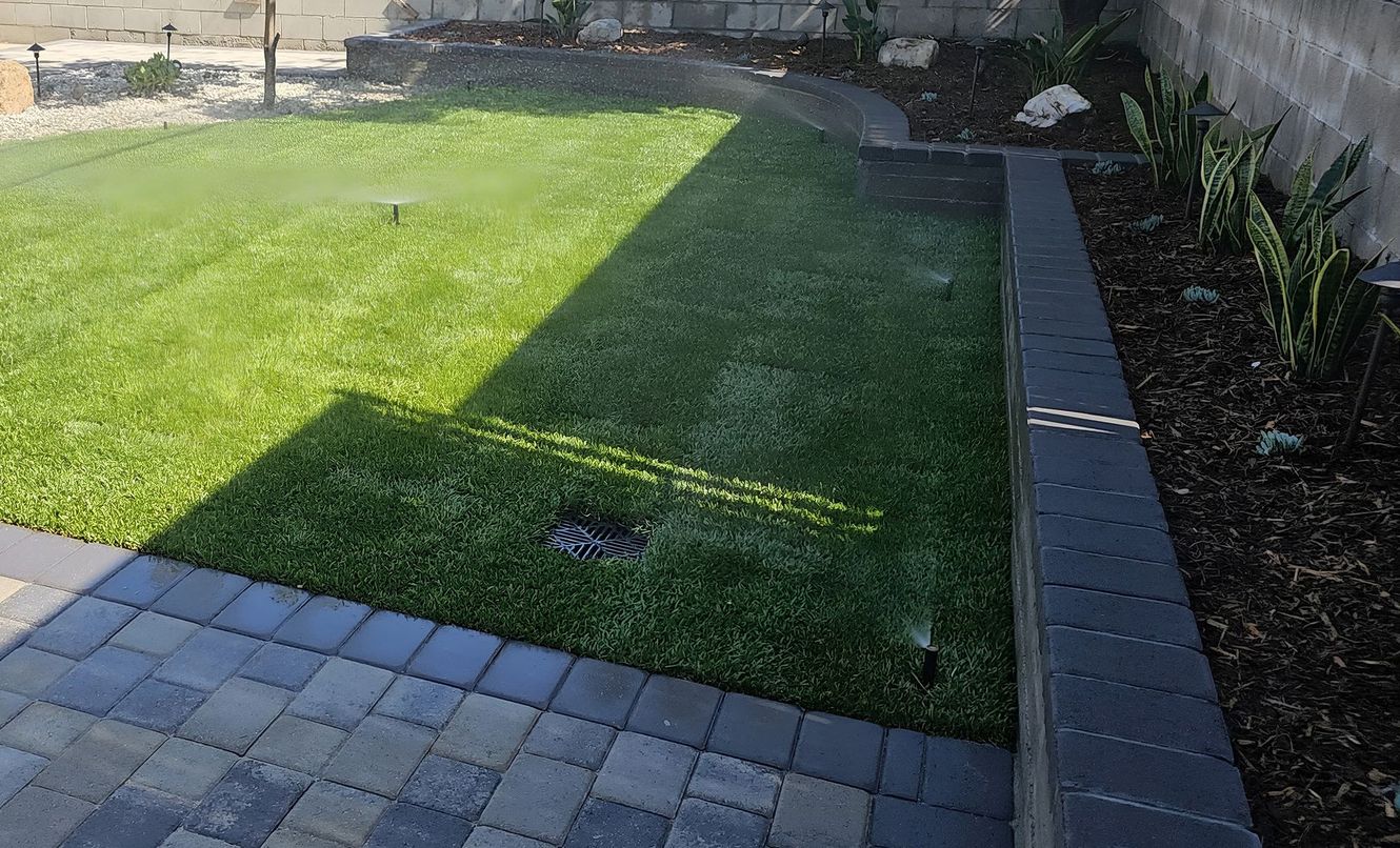 A green lawn with sprinklers, bordered by a dark-colored retaining wall and patio. Water sprays from the sprinklers.