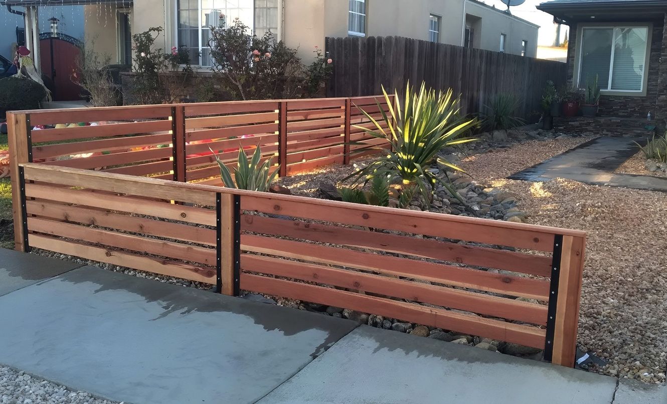 A wooden fence with horizontal planks frames a small garden bed with plants. The fence is brown, and the ground is covered in small rocks.