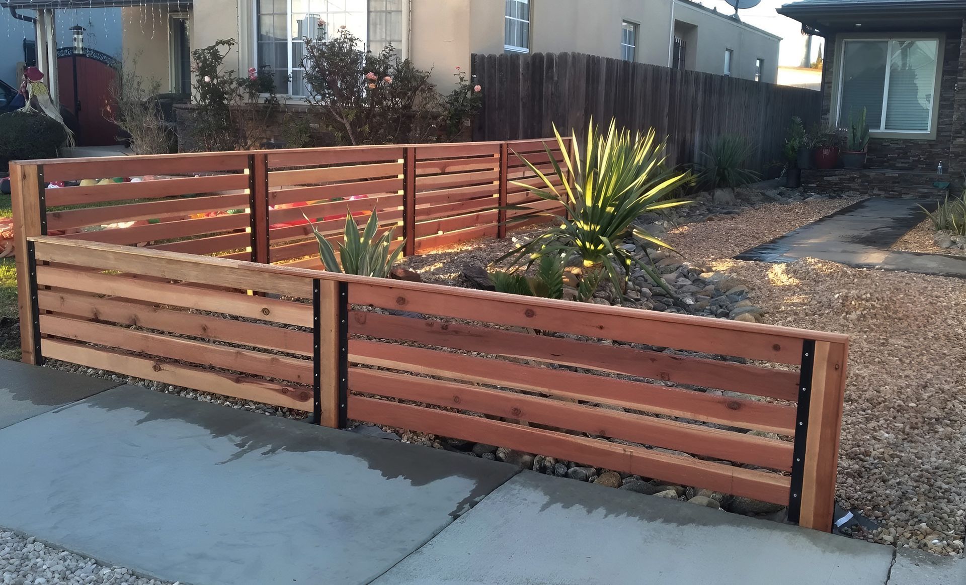 A wooden fence with horizontal planks frames a small garden bed with plants. The fence is brown, and the ground is covered in small rocks.