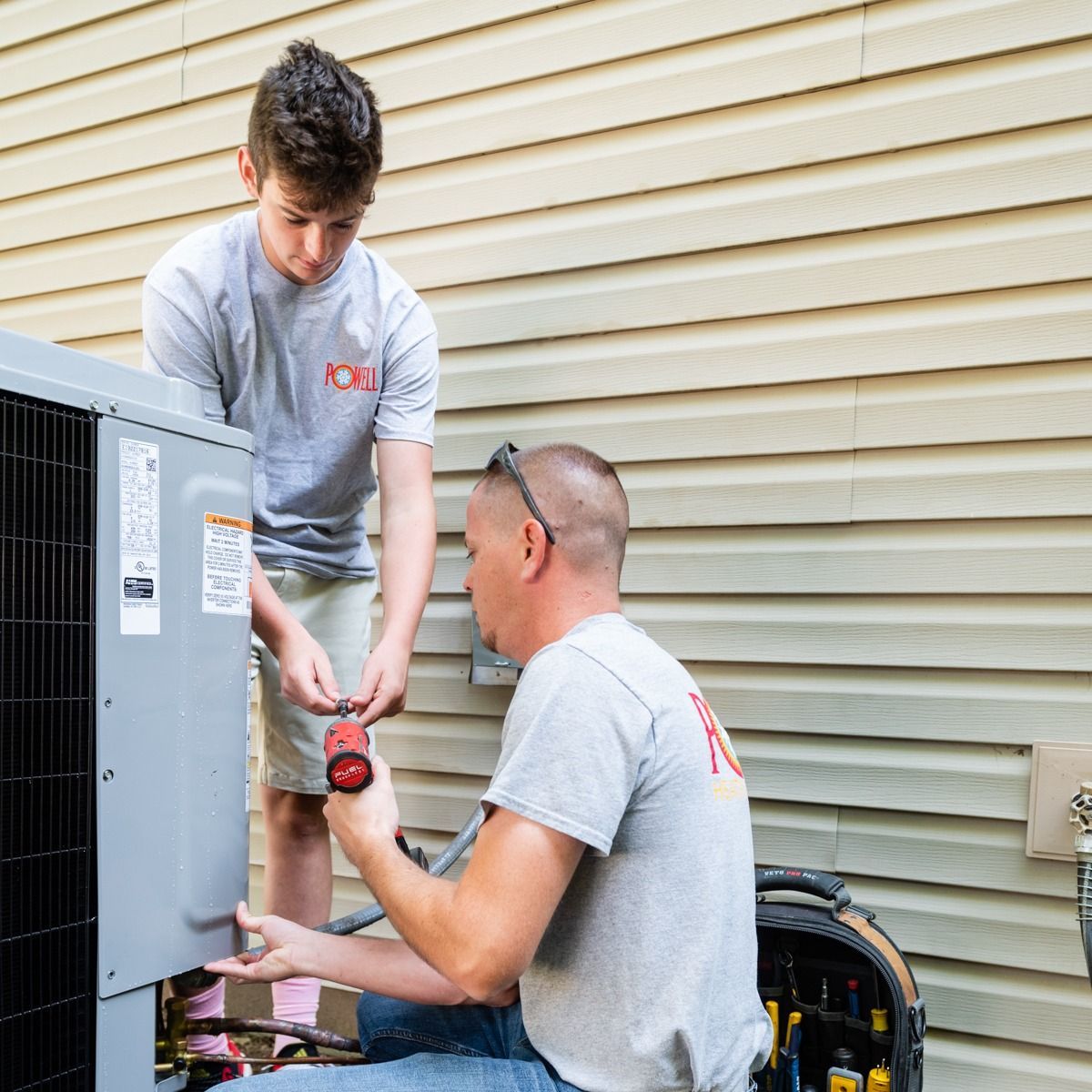 Two men are working on an air conditioner outside of a house.